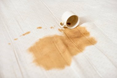 Beyaz kahve fincanı devrildi ve beyaz kumaşta kahverengi leke oluşturdu. Parlak ışıklandırma, temiz kumaş ve kahve dökülmesi arasında güçlü bir kontrastı vurguluyor. Olay yeri kaza ya da dikkatsizlik olduğunu gösteriyor.