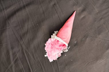 Pembe dondurma külahı karanlık yüzeyde yan yatıyordu, kısmen ezilmişti. Aydınlatma yumuşak gölgeler oluşturur, nötr gri arka planda canlı pembeyi vurgular. Beklenmedik ama neşeli anları yakalar