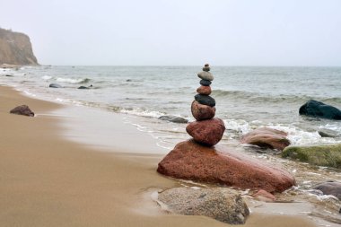 Okyanusla kumlu sahilde dengelenmiş pürüzsüz taşlar yığını. Yumuşak dalgalar yaklaşıyor, sakin bir ortam yaratıyor. Gökyüzü bulutlu ve arka plandaki kayalıklar sükuneti artırıyor