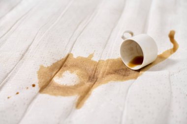 Dokulu beyaz kumaşa yakın kahve dökülmüş. Yan tarafta duran boş bardak soyut leke deseni oluşturur. Sıcak tonlar ve hafif ışıklandırma sahneyi güçlendirir