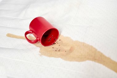 Kırmızı kupa beyaz desenli kumaşa takla atarak yayılmış bir leke yarattı. Parlak ışıklandırma, canlı kupa ve beyaz arkaplan arasındaki karşıtlığı artırarak, kaza etkisini vurguluyor