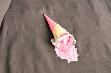 Pembe dondurma külahı karanlık yüzeyde yan yatıyordu, kısmen ezilmişti. Aydınlatma yumuşak gölgeler oluşturur, nötr gri arka planda canlı pembeyi vurgular. Beklenmedik ama neşeli anları yakalar