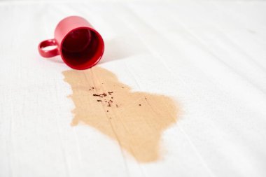 Kırmızı bardak devrilmiş, beyaz desenli yüzeye kahve dökülüyor. Kahverengi sıvı, görünür kahve telveleriyle düzensiz lekeler yaratır. Yumuşak ışık renkleri ve dokuları vurgular