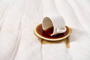 Beyaz kahve fincanı beyaz noktalı kumaşın üzerine ters yatar, fincan tabağına ve kumaşa kahve döker. Parlak ışıklandırma temiz, minimalist bir atmosfer yaratır. Eğik açı, kazara döküldüğünü vurgular.
