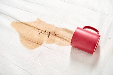 Kırmızı kupa beyaz kumaşın üzerine kahve döküyor. Hafif kahve lekesi yayılır ve sıçrama etkisi yaratır. Karşıt renkler öne çıkan lekeleri vurgular