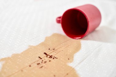 Kırmızı bardak devrildi, beyaz kumaşın üzerine kahve döküldü. Kahve taneleri yüzeye saçılmış. Parlak ışıklandırma kırmızı kupa ve beyaz arkaplan arasındaki karşıtlığı vurgular
