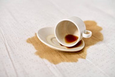 Beyaz masa örtüsünün üzerine konmuş boş kahve fincanı kahverengi sıvı leke yapıyor. Yumuşak ışıklandırma, beyaz kumaş ve koyu dökülme, kaza hissi ve sakarlık arasındaki zıtlığı vurgular.