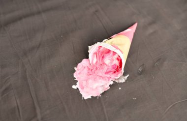 Pembe dondurma külahı karanlık yüzeyde yan yatıyordu, kısmen ezilmişti. Aydınlatma yumuşak gölgeler oluşturur, nötr gri arka planda canlı pembeyi vurgular. Beklenmedik ama neşeli anları yakalar