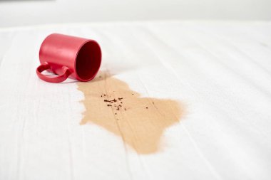 Kırmızı bardak devrilmiş, beyaz desenli yüzeye kahve dökülüyor. Kahverengi sıvı, görünür kahve telveleriyle düzensiz lekeler yaratır. Yumuşak ışık renkleri ve dokuları vurgular