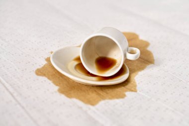 Beyaz masa örtüsünün üzerine konmuş boş kahve fincanı kahverengi sıvı leke yapıyor. Yumuşak ışıklandırma, beyaz kumaş ve koyu dökülme, kaza hissi ve sakarlık arasındaki zıtlığı vurgular.