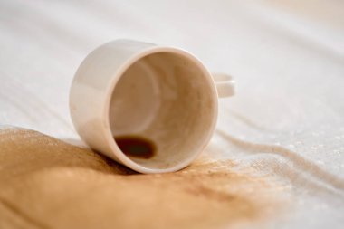 Beyaz fincanın kumaş üzerine devrilmesi kahverengi kahve lekesinin dışa doğru yayılmasıyla sonuçlandı. Işık arkaplanı koyu sıvının kontrastını arttırır. Yakın çekim açısı dağınık detayları yakalar