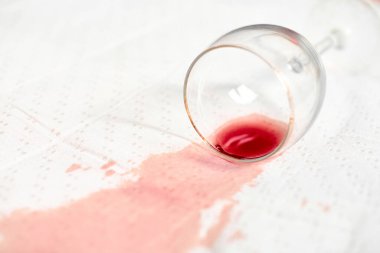 Kırmızı şarap lekesi beyaz desenli kumaşa yayılarak kontrast oluşturuyor. Boş bardaklar yan yatmış, bu da dökülmesine neden olmuş. Yumuşak ışıklandırma renk yoğunluğunu ve doku detaylarını arttırır