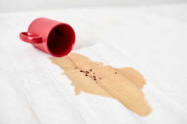 Kırmızı bardak devrildi, beyaz yüzeye kahve döküldü. Kahverengi sıvı leke yayılır ve temiz arkaplanda kontrastlar yaratır. Ekran perspektifinden çekilen sahne