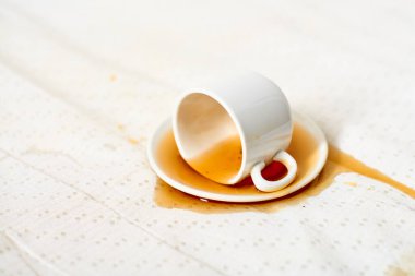 Beyaz fincan, yana yatırılmış, çay tabağının üzerinde, kahve lekeli kumaş. Yumuşak ışıklandırma, kumaş yüzeyine dökülme desenini vurgular, ince bir kontrast yaratır