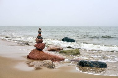 Okyanusla kumlu sahilde dengelenmiş pürüzsüz taşlar yığını. Yumuşak dalgalar yaklaşıyor, sakin bir ortam yaratıyor. Gökyüzü bulutlu ve arka plandaki kayalıklar sükuneti artırıyor