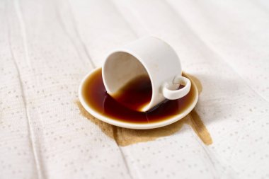 Beyaz kahve fincanı beyaz noktalı kumaşın üzerine ters yatar, fincan tabağına ve kumaşa kahve döker. Parlak ışıklandırma temiz, minimalist bir atmosfer yaratır. Eğik açı, kazara döküldüğünü vurgular.