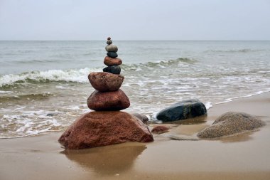 Okyanusla kumlu sahilde dengelenmiş pürüzsüz taşlar yığını. Yumuşak dalgalar yaklaşıyor, sakin bir ortam yaratıyor. Gökyüzü bulutlu ve arka plandaki kayalıklar sükuneti artırıyor