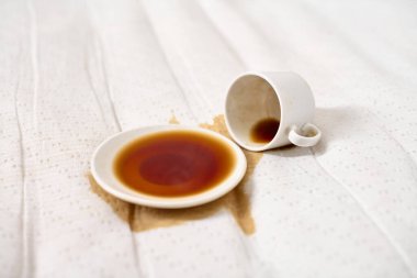 Kahve fincanı açık bej kumaşın üzerine devrilmiş. Koyu kahve çay tabağının altında havuz oluşturur. Yumuşak, dağınık ışıklandırma derindeki sıvıları vurgular