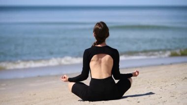 Kahverengi saçlı genç bir kadın Baltık Sahili 'nde yoga yapıyor. Meditasyonda bağdaş kurup oturuyor, siyah kıyafetler giyiyor. Sakin okyanus arkaplanı sükuneti artırır