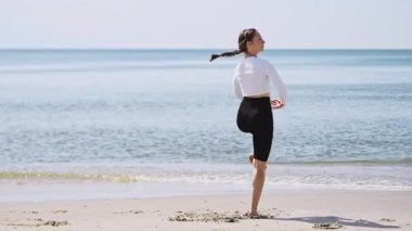 Genç yetişkin kadın, kumlu baltık sahillerinde zarif bir bale pozu veriyor. Güneşli sahne, dingin deniz ve gökyüzü arkaplanını ön plana çıkarıyor. Sakin, yansıtıcı atmosfer