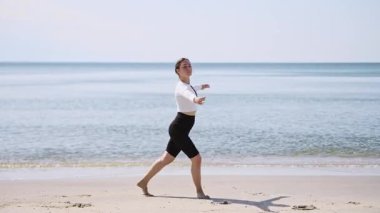 Genç bayan, Baltık Denizi 'nin arka planında kumlu sahilde bale hareketleri yapıyor. Sakin sularda güneşli bir sahne. Beyaz üst, koyu şortla tezat oluşturuyor. Zarafeti arttırıyor.