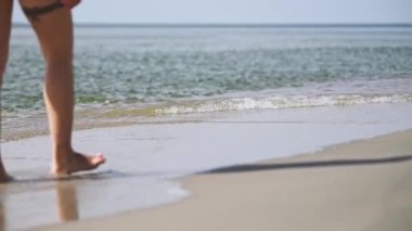 Yakından bakınca, baltık deniz kıyısında çıplak ayakla duran genç bir dişinin bacaklarını gösteriyor. Yumuşak dalgalar yumuşak yaz ışığı altında kumlu sahile dokunur ve sakin bir atmosfer yaratır.