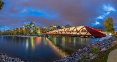 Kanada, Calgary 'deki Barış Köprüsü' nün alacakaranlıktaki panoramik görüntüsü. Kırmızı yaya köprüsü, şehrin modern silueti ve arka planında sonbahar yapraklarıyla Bow Nehri boyunca uzanır..