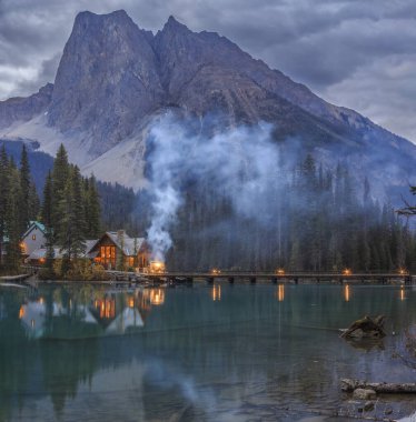 Emerald Lake in Canada, with a cozy lakeside cabin glowing at twilight, surrounded by lush forest and majestic mountains, perfectly reflecting in the calm turquoise waters. clipart