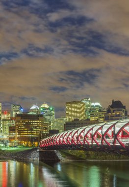 Kanada, Calgary 'deki Barış Köprüsü' nün alacakaranlıktaki panoramik görüntüsü. Kırmızı yaya köprüsü, şehrin modern silueti ve arka planında sonbahar yapraklarıyla Bow Nehri boyunca uzanır..