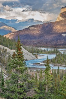 Kanada 'daki Saskatchewan Nehri' nin çarpıcı manzarası. Yoğun yeşil ormanlar ve dramatik gökyüzünün altındaki görkemli dağlarla çevrili vahşi doğanın dokunulmamış güzelliğini yakalıyor..