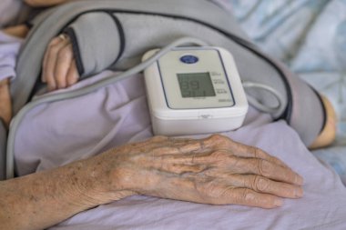 Yaşlı kişi evdeki kan basıncını dijital sphygmomanometre ile ölçüyor. Yıllanmış el ve kol manşetlerine yakın çekim, sağlık hizmetlerini, yaşlanmayı ve tıbbi yardımı yansıtıyor.