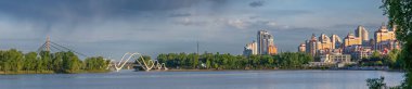 Günbatımında Kyiv 'deki Dnipro Nehri üzerindeki modern yerleşim yerleri ve yaya köprüsü. Sakin su, ağaçlar ve dramatik gökyüzü ile huzurlu bir şehir..