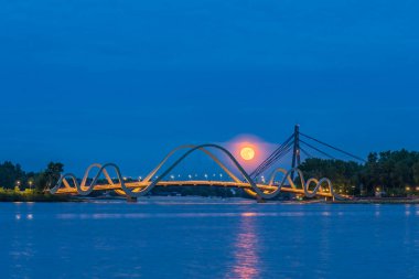 Dolunay, Kyiv 'deki Dnipro Nehri' nin üzerinde modern bir yaya köprüsünün arkasında parlayarak yükseliyor. Şehir ışıkları ve sakin su huzurlu ve rüya gibi bir akşam ortamı yaratır..