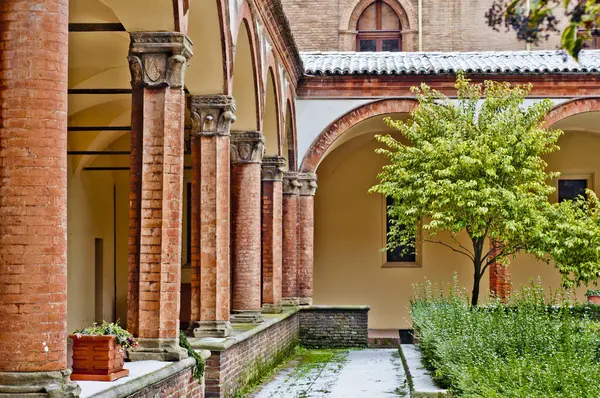 A serene courtyard within the Basilica di San Francesco in Bologna, Italy. Red brick columns, frame the open space. Lush greenery and simplicity define this principal Franciscan church