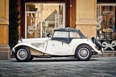 Klasik beyaz üstü açık araba kaldırım taşı bir caddeye park edilmiş, kemerli kapıları ve süslü pencereleri olan zarif bir binanın bitişiğinde. Mimaride sıcak tonlu taş maddeler ve karmaşık detaylar yer alıyor. Yumuşak ışıklandırma gölgeler oluşturur, yükseklik