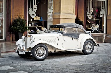 Klasik beyaz üstü açık araba kaldırım taşı bir caddeye park edilmiş, kemerli kapıları ve süslü pencereleri olan zarif bir binanın bitişiğinde. Mimaride sıcak tonlu taş maddeler ve karmaşık detaylar yer alıyor. Yumuşak ışıklandırma gölgeler oluşturur, yükseklik