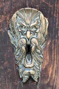 Yıpranmış ahşap bir yüzeydeki dekoratif metal kapı tokmağının yakın plan fotoğrafı. Karmaşık bir tasarım. Aslan başı motifi ve yıpranmış pirinç kaplama. Kaliteli bir his için hafif gölgeler ve toz görünür.