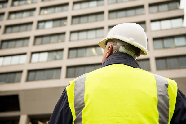 View from behind a buildings inspector or construction worker facing a building exterior, wearing a white safety helmet and yellow high visibility vest.