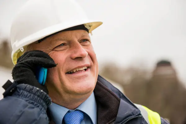 Portrait of a businessman buildings inspector or construction worker, using a mobile phone, and wearing a white safety helmet with yellow high visibility vest.