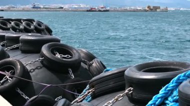 Cebelitarık 'taki rıhtım ekipmanlarının geniş açılı görüntüsü. Havada süzülen lastik çamurluklar güçlü rüzgar ve dalgalarla hareket ettirilir. Körfezin üstünde beyaz bulutlu mavi bir gökyüzü var..