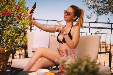 Genç gülümseyen bikinili kadın portresi otelin terasında akıllı telefonuyla selfie çekiyor. Yaşam biçimi.