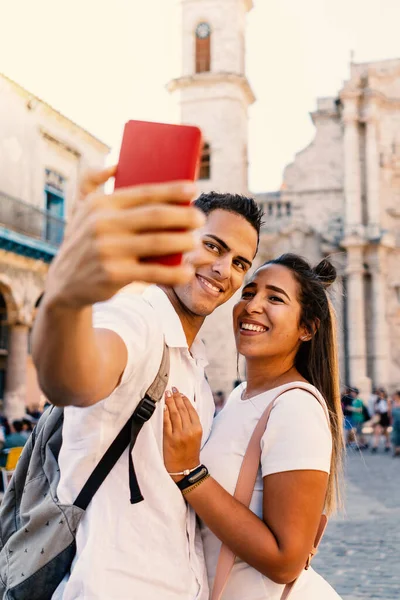 在古巴哈瓦那 一对笑着带着智能手机自拍的年轻夫妇 背景上的大教堂专注于这对夫妻 — 图库照片