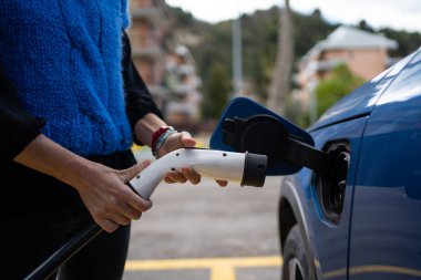Sokakta bir kadın elektrikli arabayı şarj ediyor. Çevre hareketliliği kavramı. Elektrikli araca bağlı güç kaynağı.