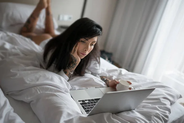 官能的な若い女性は 朝にコーヒーのクオを飲みながら 彼女のベッドに横たわって ポータブルコンピュータを使用して肖像画を閉じます — ストック写真