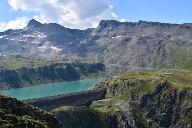 Görüntü, dağlarda yuva yapmış Diga del Goillet barajının engebeli kayalık arazilerle çevrili büyük bir turkuaz rezervuarının panoramik manzarasını gösteriyor. Arkaplanda karla kaplı zirveler yükseliyor