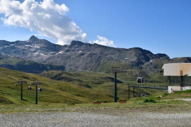 Vadide hareket eden teleferikli çarpıcı bir dağ manzarası. Yeşil çayırlar ve karla kaplı tepeler açık mavi gökyüzünün altında nefes kesici bir zemin oluşturur. Köşede bir teleferik durağı var.