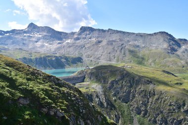 The image shows a panoramic view of the Diga del Goillet lake dam nestled in the mountains, with a large turquoise reservoir surrounded by rugged, rocky terrain. Snow-capped peaks rise in the clipart