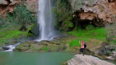 Kadın elinde bir cep telefonu tutuyor ve güzel bir şelalenin fotoğrafını çekiyor. Yavaş çekim videosu. 