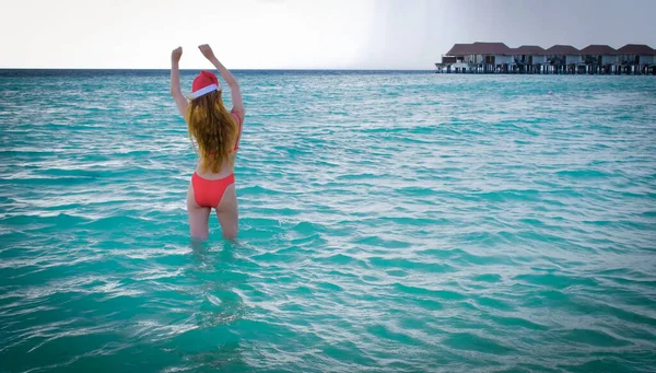 Woman from a back with a long hair, red bikini and in a Santa hat. Blue turquoise ocean on the background. Girl enjoys her tropical holidays on Maldives. Summer travel vacation concept.