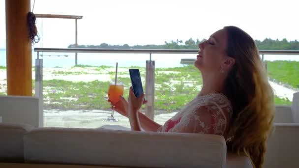핸드폰을 칵테일을 마시는 몰디브의 잔하는 청록색 배경에 있습니다 소녀는 휴일을 — 비디오